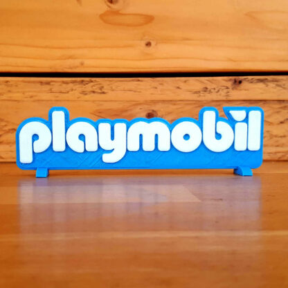 Logo 3D Playmobil bleu posé sur son socle, sur un plan de travail en bois